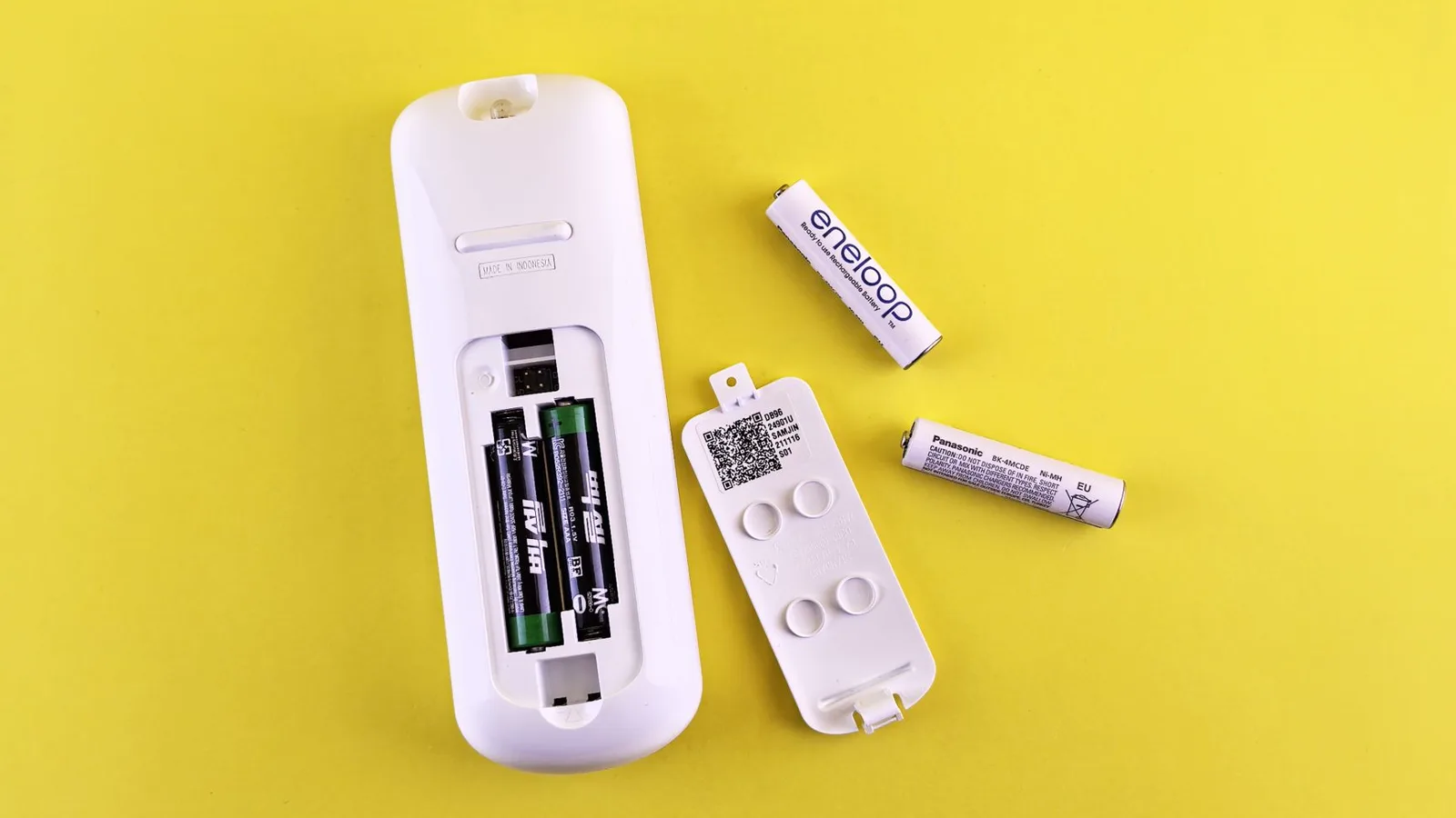Pilha alcalina descartável (prata) e pilha recarregável NiMH (dourada) lado a lado em mesa de madeira, com controle remoto desfocado ao fundo e carregador USB visível na borda, luz natural de janela em interior doméstico