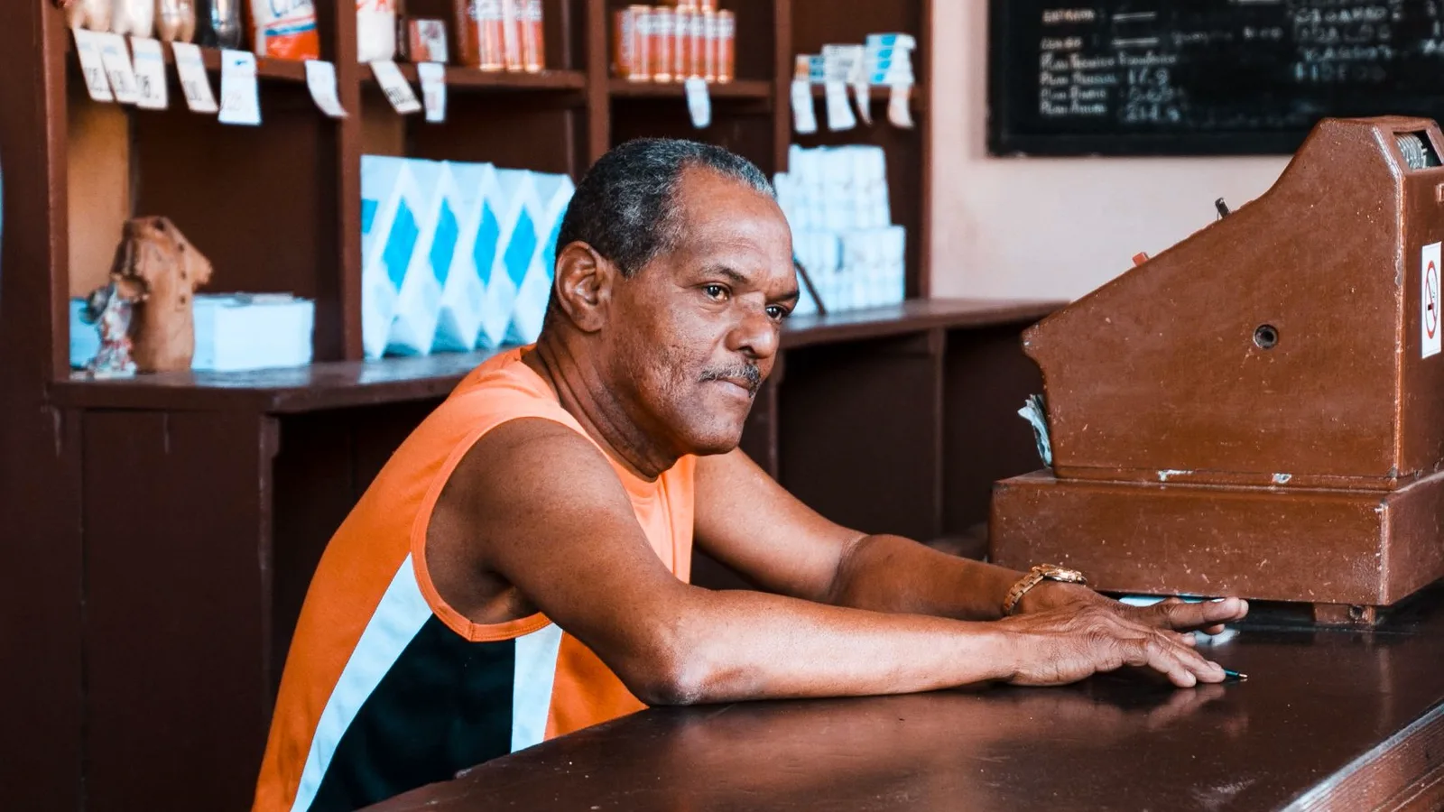 Lojista conferindo a etiqueta traseira de computador desktop em balcão de comércio, com UPS branca ao lado e tomada à vista, sugerindo planejamento de proteção elétrica.