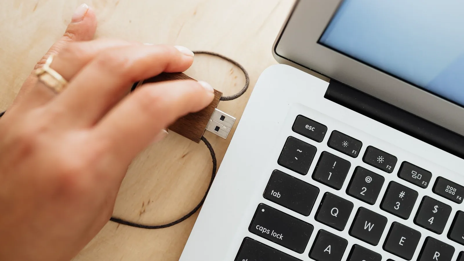 Mão inserindo pen drive em porta USB azul de computador em ambiente de trabalho doméstico, com outro pen drive e cabos ao fundo fora de foco.
